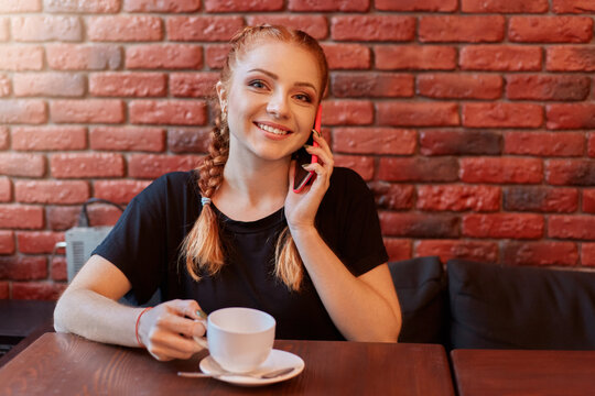 Attractive girl talking on phone while drinking coffee in cozy cafe against brick wall, smiling red haired lady spending free time in cafeteria, wearing black t shirt. - Powered by Adobe