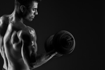Muscular model sports young man with dumbbells in hand on dark background. Black and white fashion portrait of strong brutal guy. Sexy torso. Male flexing his muscles.