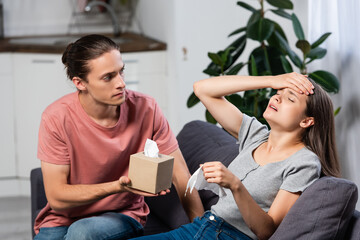 young man holding paper napkins near ill girlfriend suffering from migraine with closed eyes