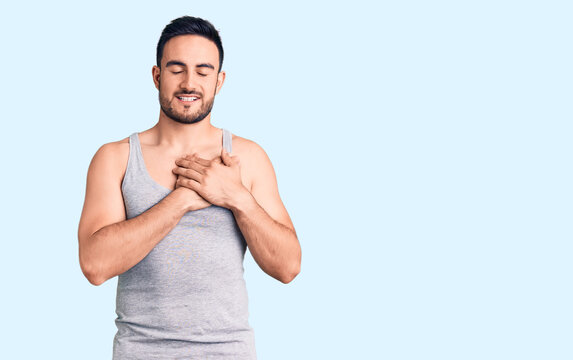 Young handsome man wearing swimwear and sleeveless t-shirt smiling with hands on chest with closed eyes and grateful gesture on face. health concept.