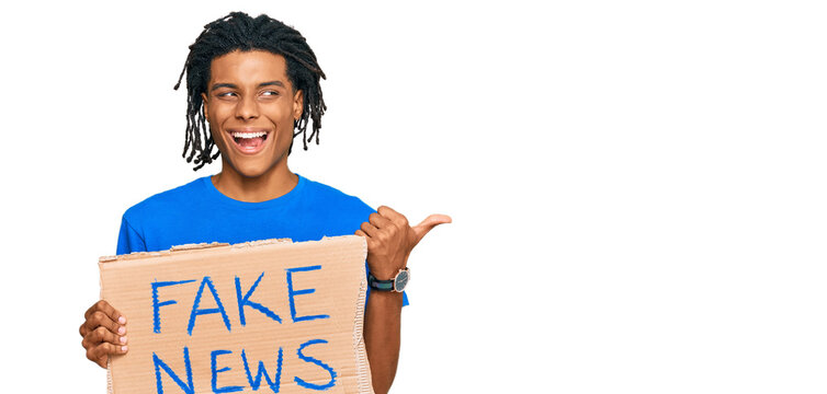 Young african american man holding fake news banner pointing thumb up to the side smiling happy with open mouth