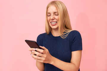 Naklejka premium Beautiful emotional young European woman with teeth brackets and freckles chatting online with friends via application on mobile. Funny teenage girl watching videos on smart phone, making grimace