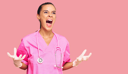 Young hispanic woman wearing doctor uniform and stethoscope crazy and mad shouting and yelling with...