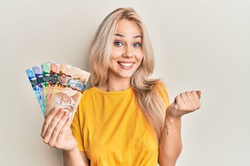 Beautiful caucasian blonde girl holding canadian dollars screaming proud, celebrating victory and success very excited with raised arm