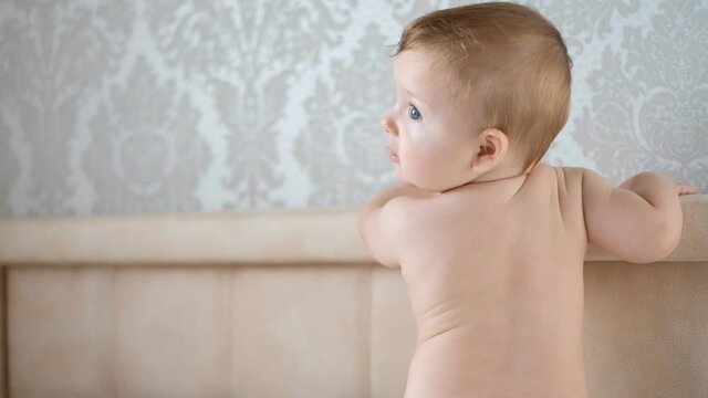 A kid without clothes stands on his parents' bed in the morning. A child without clothes walks on the bed.