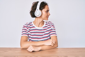 Young hispanic woman using headphones sitting on the table looking to side, relax profile pose with natural face with confident smile.