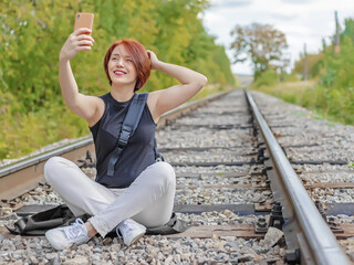 happy pretty red-haired young woman sitting on the railroad track with phone in her hands