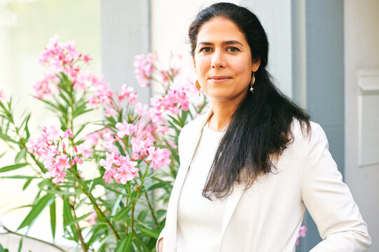 Beautiful Middle Age Woman Posing Outside, Wearing White Jacket, Standing Next Too Pink Flowers