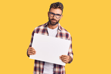 Young hispanic man holding blank empty banner thinking attitude and sober expression looking self confident