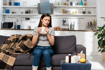 sick woman holding paper napkin while sitting on sofa near plaid blanket and bedside table with medicines