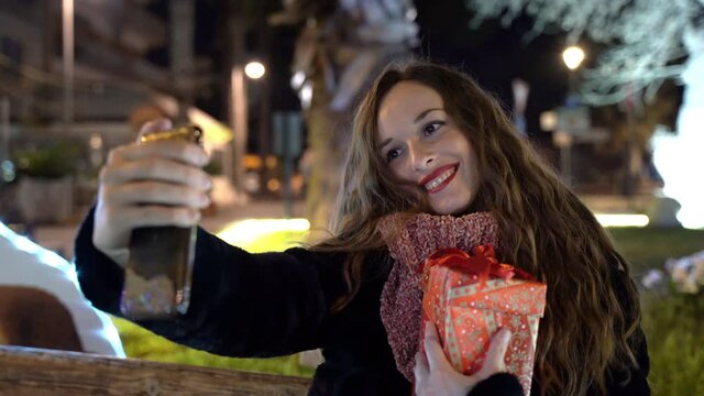 Pretty woman with christmas present in hands using smartphone and taking selfie photos in the park at night, amazing illuminated street with christmas lights and garlands, holidays and christmas