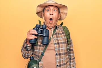 Senior man with grey hair wearing hiker bakcpack holding binoculars scared and amazed with open mouth for surprise, disbelief face