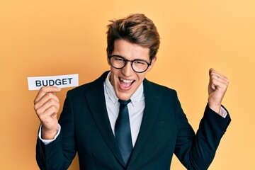 Handsome caucasian business man holding budget word on paper screaming proud, celebrating victory and success very excited with raised arms