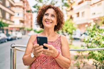 Middle age woman smiling happy using smartphone walking at street of city.