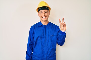 Young hispanic boy wearing worker uniform and hardhat smiling with happy face winking at the camera doing victory sign. number two.
