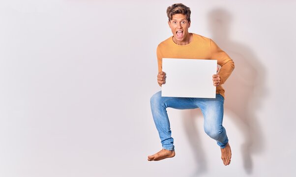 Young handsome blond man with open mouth. Holding empty blank banner jumping over isolated white background
