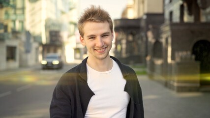 Fototapeta premium Young smiling guy dressed casually on the background of city road. Middle shot of smiling man standing near the road, on background of blurred residential houses