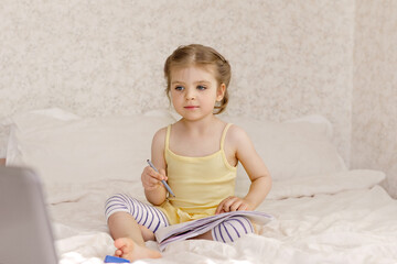 girl 3 - 4 years Caucasian in yellow dress sits in bedroom of white bed in front of laptop and draws in album concept of home education,