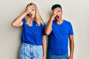 Young interracial couple wearing casual clothes peeking in shock covering face and eyes with hand, looking through fingers with embarrassed expression.
