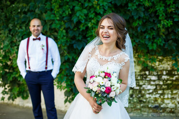 The bride in a white dress in the foreground and the groom with suspenders stands behind. on the background wall with green leaves