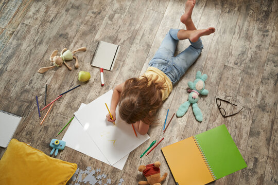 Top view of little caucasian girl lying on the wooden floor at home and drawing with colorful pencils on a white sheet of paper
