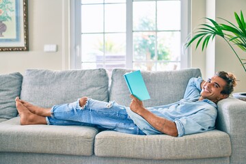 Young hispanic man smiling happy reading book laying on the sofa at home