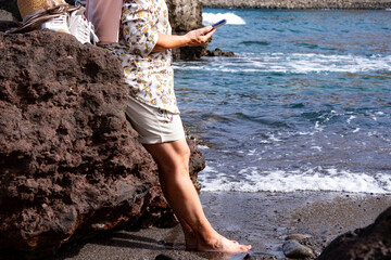Senior woman using smart phone standing on the rocks with feet into the fresh water enjoying the vacation at sea - concept of active elderly during retirement