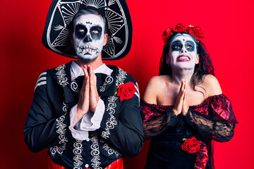 Young couple wearing mexican day of the dead costume over red begging and praying with hands together with hope expression on face very emotional and worried. begging.