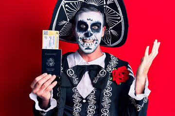 Young man wearing mexican day of the dead costume holding united states passport and boarding pass celebrating victory with happy smile and winner expression with raised hands