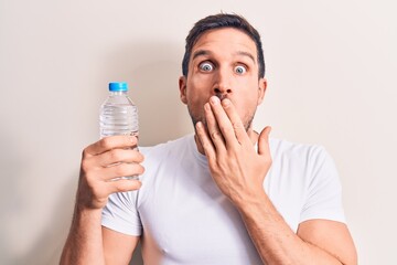 Young handsome man drinking bottle of water to refreshment over isolated white background covering mouth with hand, shocked and afraid for mistake. Surprised expression