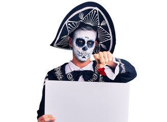 Young man wearing day of the dead costume holding blank empty banner pointing with finger to the camera and to you, confident gesture looking serious