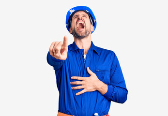 Young handsome man wearing worker uniform and hardhat laughing at you, pointing finger to the camera with hand over body, shame expression