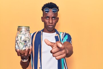 Young african american man holding jar with savings pointing with finger to the camera and to you, confident gesture looking serious