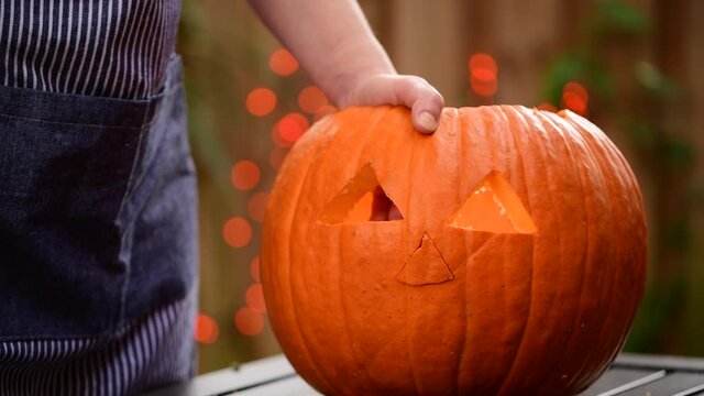 Carved pumpkin on Halloween. Woman hands make a Jack O Lantern face decoration from fresh pumpkins. Autumn Season. Scary Jack-o'-Lantern face. Orange lights Bokeh on background. 4K