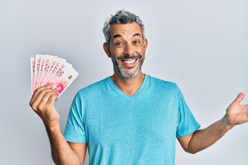 Middle age grey-haired man holding israel shekels celebrating achievement with happy smile and winner expression with raised hand