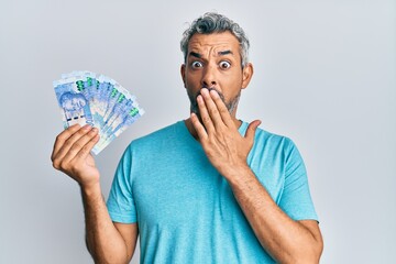 Middle age grey-haired man holding south african rands banknotes covering mouth with hand, shocked and afraid for mistake. surprised expression