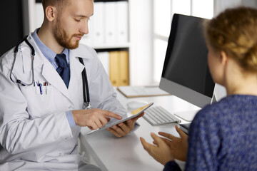 Friendly red-bearded doctor and patient woman discussing current health examination while sitting and using tablet computer in clinic. Medicine concept