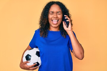 Young african american woman holding football ball talking on the smartphone sticking tongue out happy with funny expression.