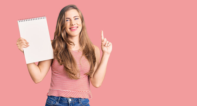 Beautiful caucasian young woman holding notebook surprised with an idea or question pointing finger with happy face, number one