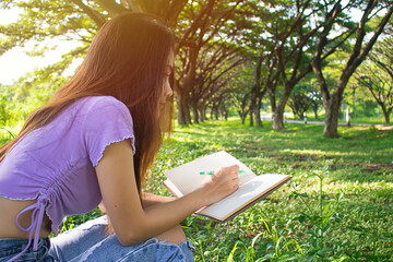 Young woman sitting thinking something on grass below tree in park. Hands holding notebook and pen for write something on nice day. Copy space.