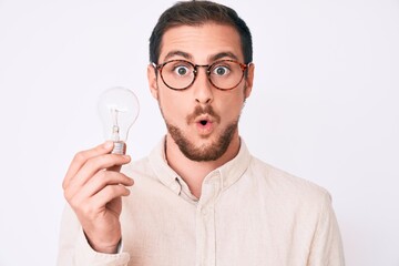 Young handsome man holding lightbulb for inspiration and idea scared and amazed with open mouth for surprise, disbelief face