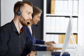 Red-bearded businessman talking by headset near his female colleague while sitting in modern...