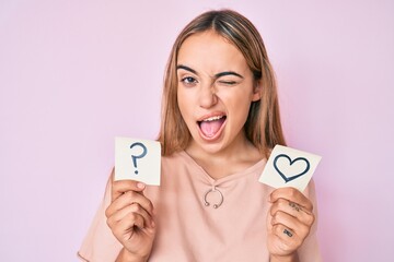 Young beautiful blonde woman holding heart and question mark reminder winking looking at the camera...
