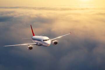 Naklejka na ściany i meble Airplane is flying above the clouds at sunset in summer. Landscape with passenger airplane, low clouds, orange sky. Front view of aircraft. Business travel. Commercial plane. Aerial view. Take off