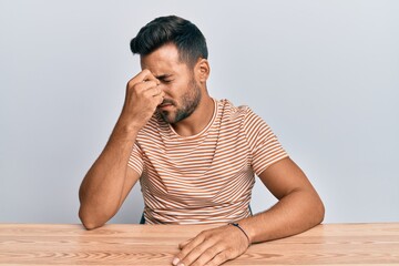 Handsome hispanic man wearing casual clothes sitting on the table tired rubbing nose and eyes feeling fatigue and headache. stress and frustration concept.