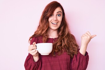 Young beautiful woman holding coffee pointing thumb up to the side smiling happy with open mouth