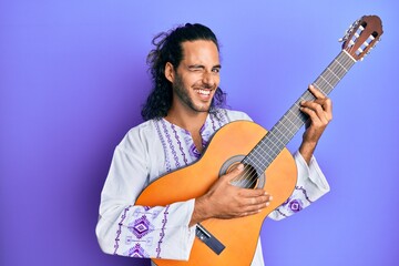 Young handsome man with long hair playing classical guitar winking looking at the camera with sexy...