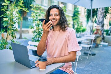 Young hispanic man smiling happy working using laptop and talking on thesmartphone at terrace of coffee shop.