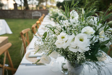 Beautiful decoration of a wedding banquet in the green garden in white colors. Serving the table with white tablecloth, plates, glasses, cutlery, napkins decoration, with white flowers in vase