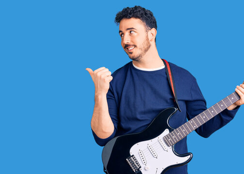 Young hispanic man playing electric guitar pointing thumb up to the side smiling happy with open mouth